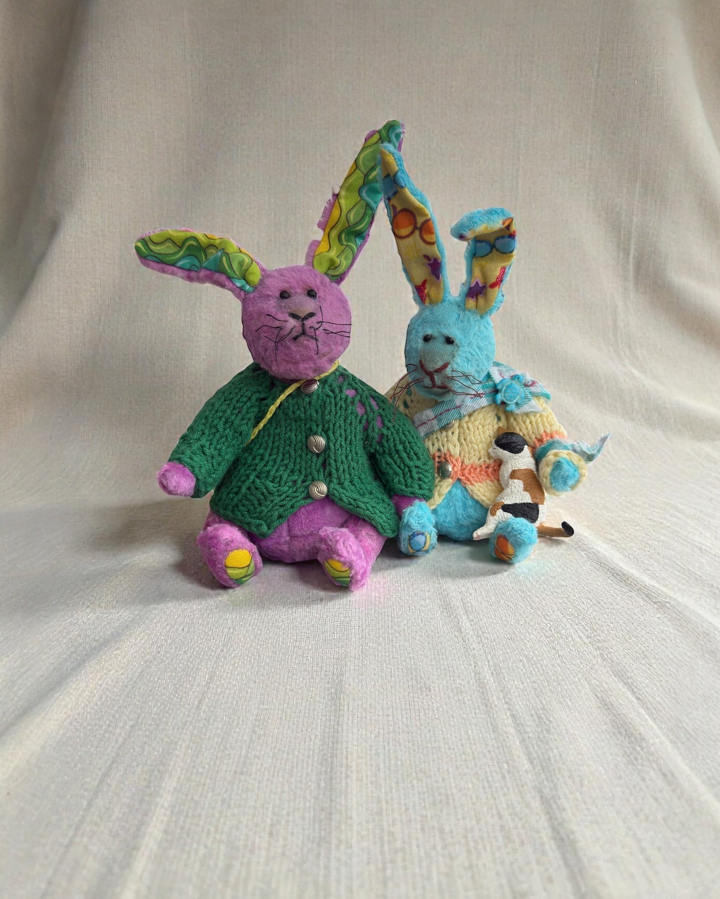 Two handmade plush bunny brothers sitting together on a soft linen backdrop, wearing knitted sweaters and showing their contrasting brave and shy personalities.