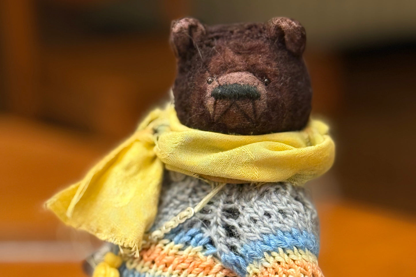 Small teddy bear wearing a colorful sweater and yellow scarf on a wooden surface